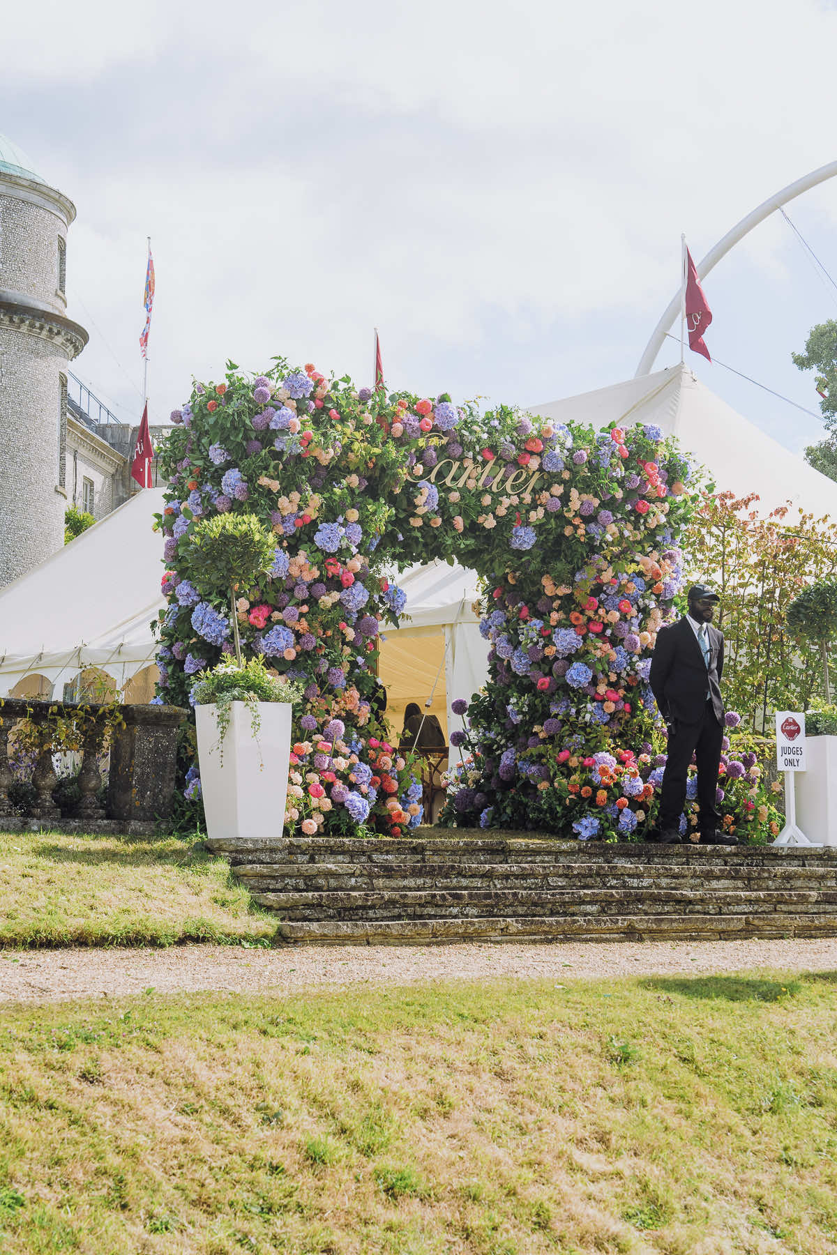 A large, flower-adorned archway made by Blooming Haus at the entrance of a white event tent. The arch is covered in colorful hydrangeas and roses. A suited man stands near a sign reading "Cartier." The tent is set up in front of a historic building with multiple red flags flying in the background.