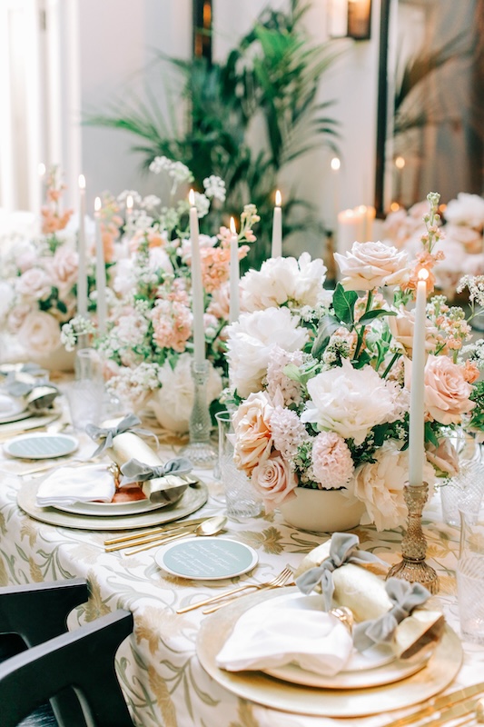 An elegantly set dinner table is adorned with tall lit candles and lush floral centerpieces containing pink and white wedding flowers from a renowned London florist. Each place setting features a white napkin with a grey ribbon, gold and white plates, and gold cutlery. The background is softly blurred greenery and a large mirror.