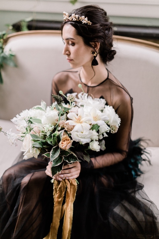 A woman in a black, sheer dress sits on a couch, holding a large bouquet of white flowers accented with greenery and beige roses from a luxury London florist. Her dark hair is styled in an updo, adorned with a delicate golden hairpiece. She gazes downwards, giving the image a serene and contemplative feel.
